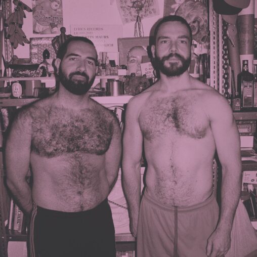 Black and white photo of two shirtless men, both are fit, bearded, and hairy chested, and are standing against a wall of posters and other ephemera.